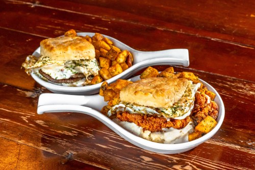 Two plates of hearty breakfast sandwiches with biscuits, fried chicken, eggs, and seasoned potato cubes on a wooden table.