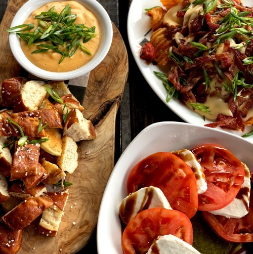 A wooden platter with pretzel bites, a bowl of cheese dip, and a plate of loaded fries topped with bacon and green onions.