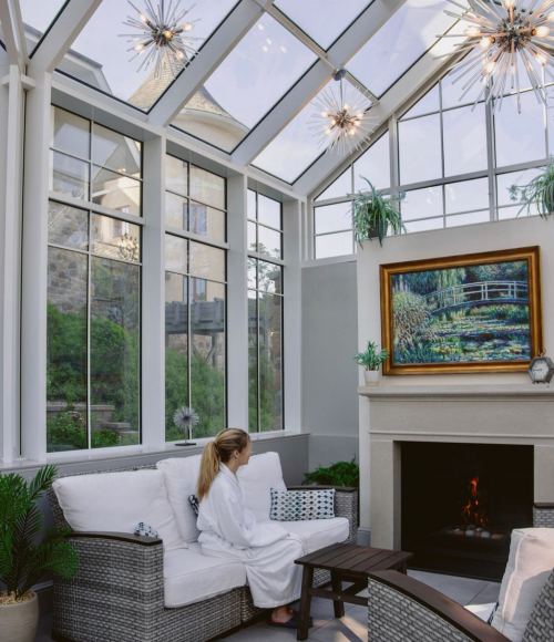 A woman in a robe sits in a bright sunroom with large windows, a fireplace, and a painting of a serene landscape.