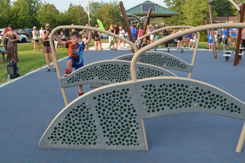 A young boy climbs on a playground structure while a crowd enjoys the park in the background.