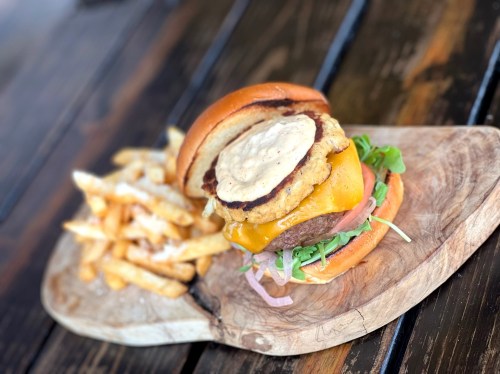 A close-up of a burger with cheese, fried chicken, and toppings, served with a side of crispy fries on a wooden board.