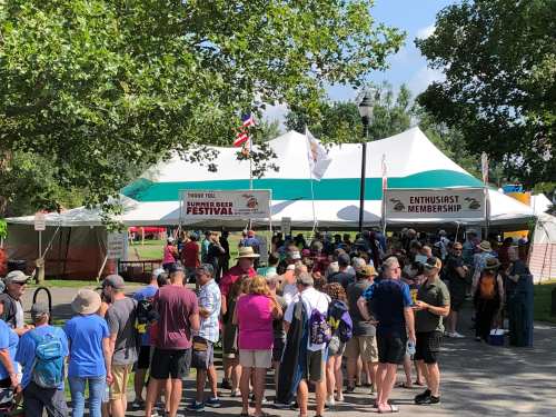 A crowd gathers outside a large tent for a summer beer festival, with banners and trees in the background.
