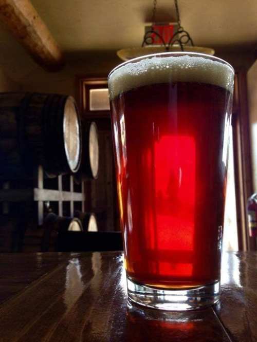 A glass of amber beer sits on a wooden table, with barrels in the background and soft lighting.