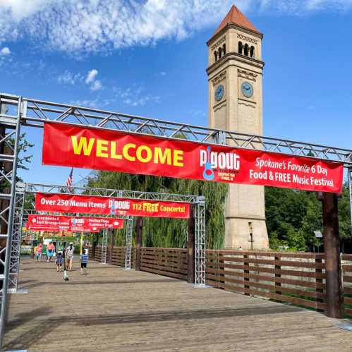 A welcome banner for a food and music festival in Spokane, with a clock tower in the background and people walking on a bridge.