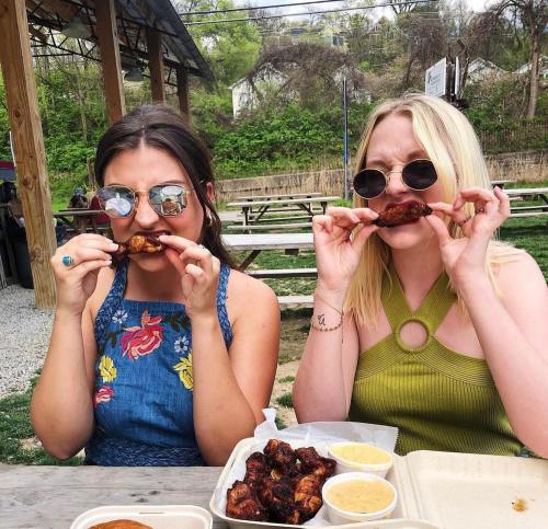 Two women wearing sunglasses playfully hold chicken wings to their mouths, posing outdoors at a picnic table.