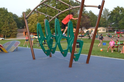A child climbs on a green playground structure while others play in the background on a sunny day.