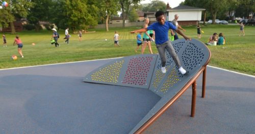 A child plays on a colorful climbing structure in a park, with other kids and activities in the background.