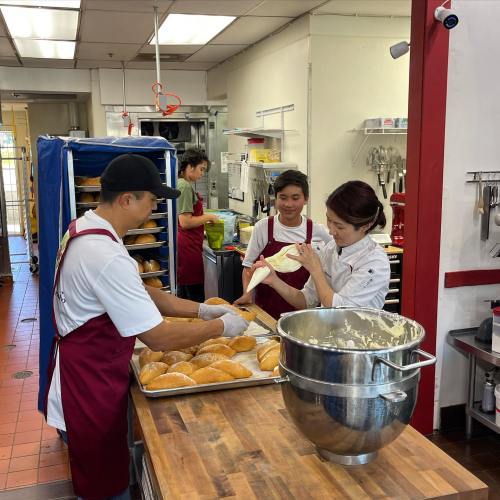 Three bakers in a kitchen prepare bread, with one mixing dough and two arranging baked goods on a table.