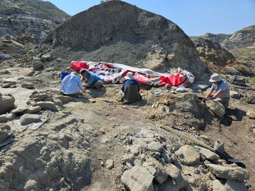 Two people work on a rocky excavation site, with a large red and white tarp in the background and hills in the distance.
