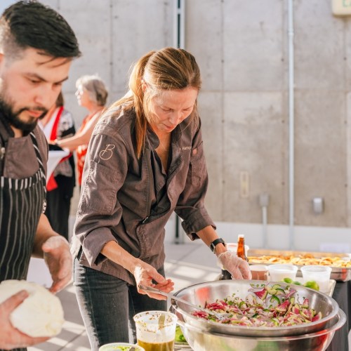 A chef prepares a salad in a large bowl, while another person assists in a vibrant kitchen setting.