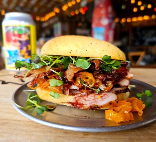 A close-up of a sandwich with layers of meat, greens, and a side of orange slices on a metal plate.