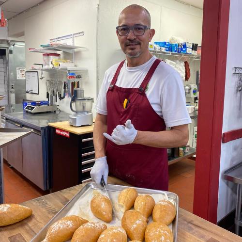 A smiling baker in an apron prepares dough on a tray in a kitchen, wearing gloves and standing by a work surface.
