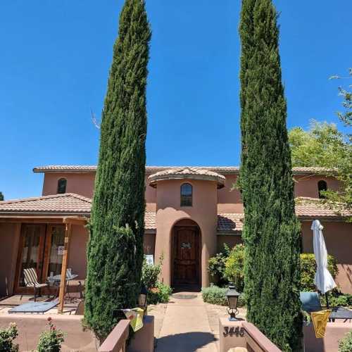 A charming two-story house with tall cypress trees, a wooden door, and a patio under a clear blue sky.