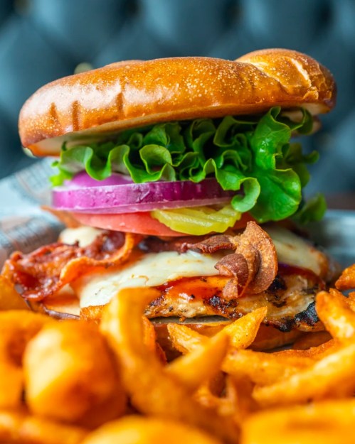 A close-up of a juicy burger with lettuce, onion, pickles, and bacon, served with curly fries on the side.