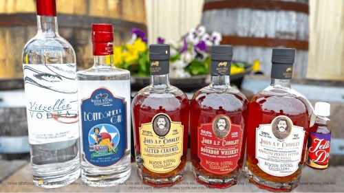 A lineup of various bottles of vodka and bourbon in front of wooden barrels and colorful flowers.