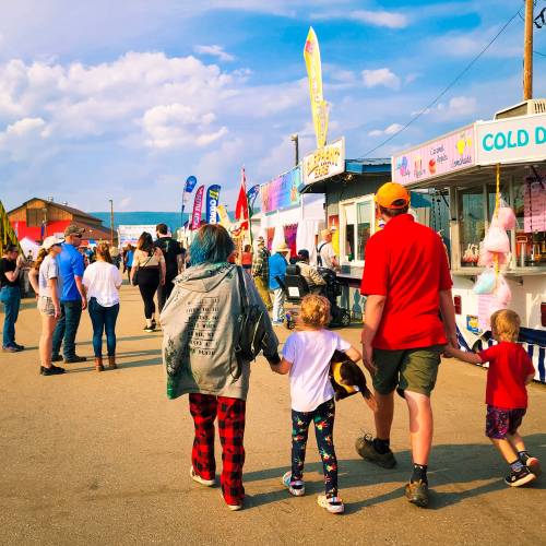 A family walks hand-in-hand through a lively fairground with colorful booths and banners under a blue sky.