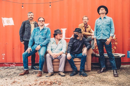 A six-member band poses on a couch outdoors, with a colorful backdrop and casual attire, exuding a relaxed vibe.