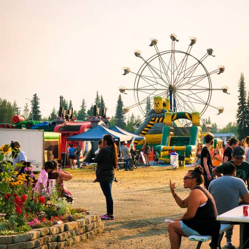 A lively fair scene with a Ferris wheel, inflatable attractions, and people enjoying the festivities amidst greenery.