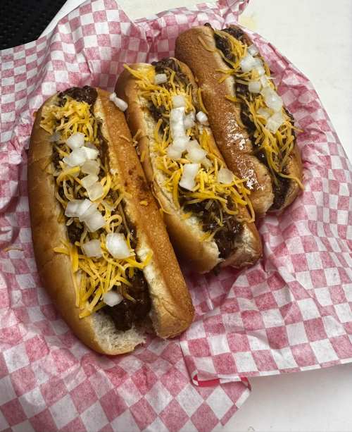 Three hot dogs topped with chili, shredded cheese, and diced onions, served on a red and white checkered paper.