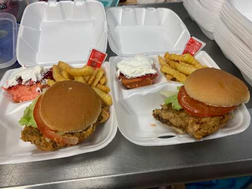 Two takeout containers with fried chicken sandwiches, lettuce, tomato, and crinkle-cut fries on the side.