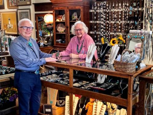 Two smiling individuals stand behind a jewelry display, surrounded by various necklaces and accessories in a shop.