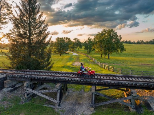 Two people ride ATVs across a wooden railway bridge, surrounded by lush green fields and trees at sunset.