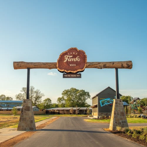 A wooden archway sign reading "Camp Fimfo Waco" welcomes visitors, set against a clear blue sky.