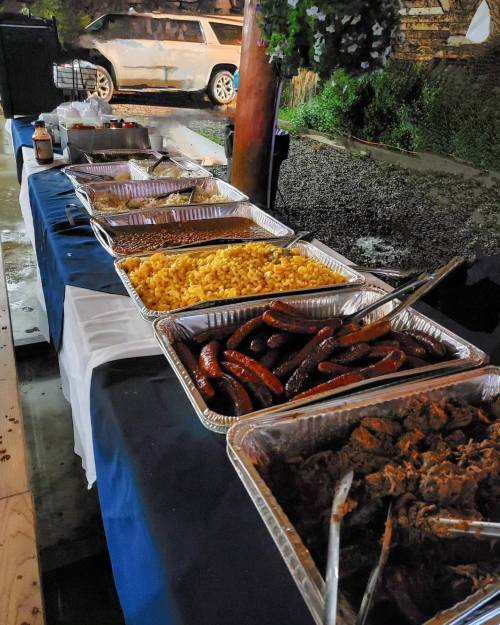 A buffet table outdoors featuring trays of macaroni and cheese, sausages, and meat dishes under dim lighting.