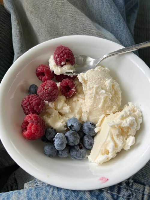 A bowl of vanilla ice cream topped with raspberries and blueberries, with a spoon resting on the side.