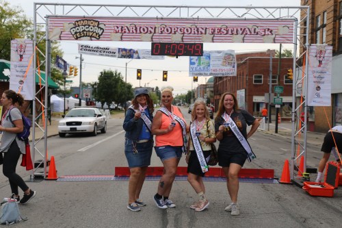 Four women in sashes pose at the finish line of the Marion Popcorn Festival 5K, with a timer showing 1:09:49.