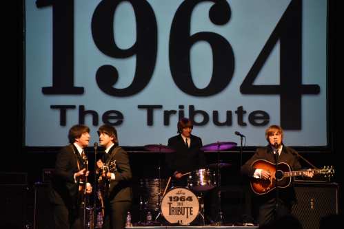 A tribute band performs on stage, dressed in suits, with a large backdrop displaying "1964 The Tribute."