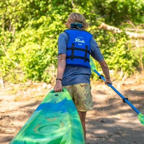 A person in a blue life jacket carries a colorful kayak and paddle along a sandy path surrounded by greenery.