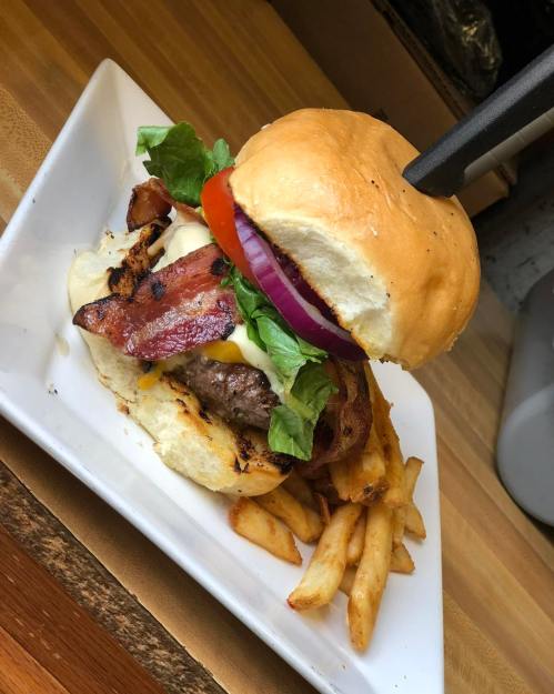 A juicy burger with bacon, lettuce, tomato, and onion, served with crispy fries on a white plate.