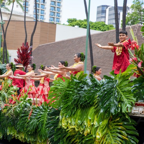 A group of dancers in traditional attire perform on a float, surrounded by greenery and city buildings in the background.