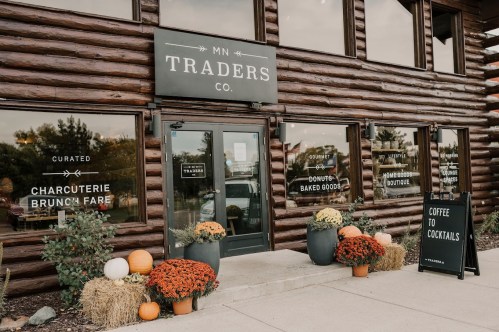 A rustic log building with a welcoming entrance, featuring signs for charcuterie, brunch, and coffee, adorned with fall decor.
