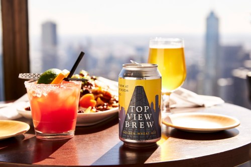 A can of Top View Brew next to a cocktail and a plate of food, with a city skyline in the background.