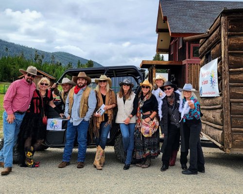 A group of people in cowboy attire poses by an off-road vehicle near a log cabin, smiling and holding brochures.