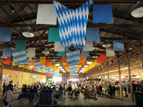 A bustling indoor festival space with colorful banners and tables filled with people enjoying food and drinks.