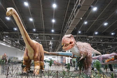 Two large dinosaur models, a brachiosaurus and a T. rex, displayed in a spacious indoor exhibition area.