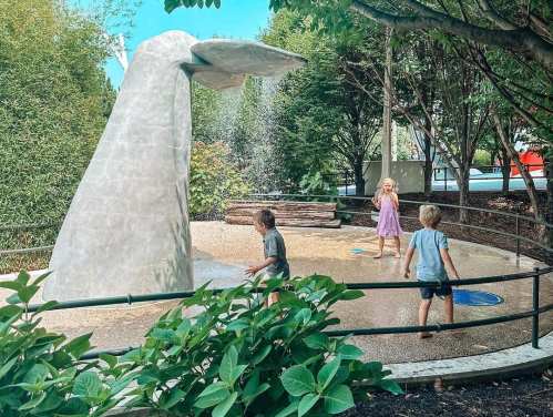 Children play in a splash pad area with a large whale sculpture surrounded by greenery.