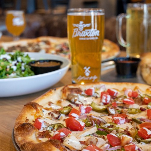 A close-up of a pizza topped with chicken, tomatoes, and peppers, with a salad and beer in the background.