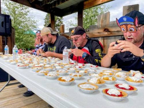 Four participants compete in a pie-eating contest, surrounded by mini pies topped with whipped cream on a table.