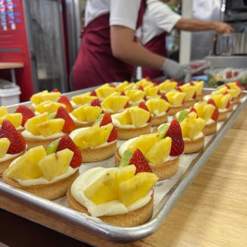 A tray of fruit tarts topped with pineapple and strawberries, set in a kitchen with a person preparing food in the background.