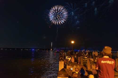 A crowd watches fireworks illuminate the night sky over a lake, with reflections on the water and a lamppost nearby.