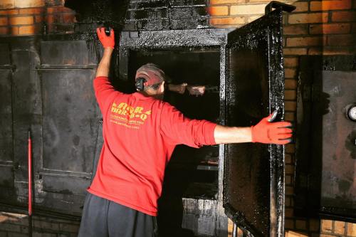 A person in a red shirt and gloves opens a large, black smoker, revealing smoke and cooking meat inside.