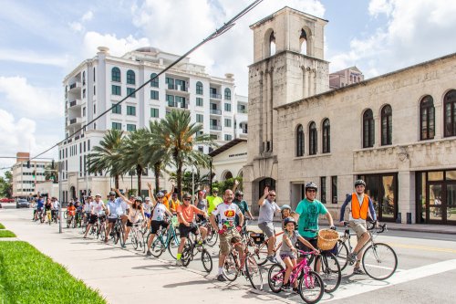 A group of cyclists in bright clothing rides along a city street lined with palm trees and buildings.
