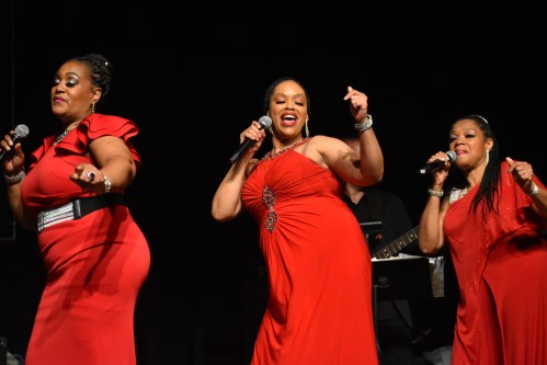 Three women in red dresses perform on stage, singing and dancing with microphones, accompanied by a musician in the background.