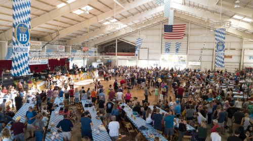 A large crowd gathers in a spacious indoor venue for a festival, with tables and a stage in the background.