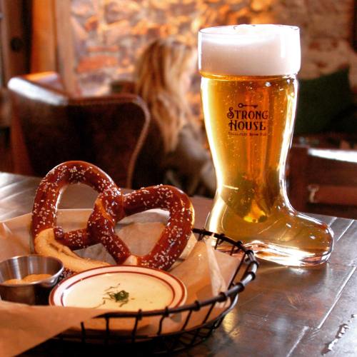 A large beer boot next to a basket of pretzels with mustard on a wooden table in a cozy setting.