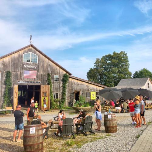 A rustic barn with an American flag, surrounded by people socializing and enjoying outdoor seating on a sunny day.